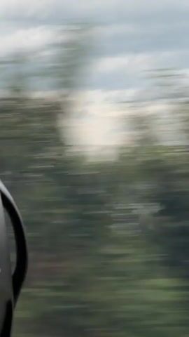 The video begins with a close-up of a helmet, partially visible on the left side of the frame. The background is blurred with motion, indicating rapid movement. Green foliage and a cloudy sky are discernible, suggesting an outdoor setting such as a road or trail. The camera appears to be mounted on a moving object, likely a bicycle or motorcycle, as suggested by the helmet. The video is brief and shows no other people or specific actions beyond the implied motion.