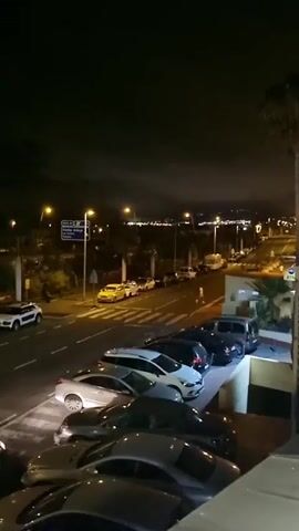 This is a nighttime, elevated view of a street scene in Miraverde, Spain. The primary subjects are several cars parked and in motion. The setting is a public road with a parking area on the right. Streetlights illuminate the scene.

The activity captured is typical urban nighttime traffic and parking. Cars are driving on the road and parked in designated spots. Some vehicles are white, others are dark (black or dark blue), and there are a few yellow cars visible in the distance.

Notable details include the time of day, which is clearly night as evidenced by the darkness and artificial lighting. The weather appears to be clear, with no rain or strong winds suggested. Location cues are limited to the text on a blue street sign visible in the distance, which reads "A-404." There are no discernible emotions conveyed by the scene, as it is a straightforward depiction of an urban environment at night. The scene is somewhat blurry due to low light conditions and possibly camera movement.