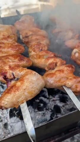 Chicken wings and meat skewers are grilling over charcoal. Smoke billows up from the grill. The chicken wings appear golden brown and are skewered on metal rods. The meat skewers have chunks of reddish-brown meat threaded onto metal rods. The scene is outdoors, with natural light filtering in. The focus is on the food being cooked.