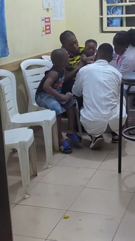 The video shows an indoor scene. In the initial frame, there are several individuals including a woman holding a crying baby, a child sitting on a plastic chair, and two people dressed in medical attire, possibly a doctor and a nurse, interacting with the woman and baby. The child has a cell phone in their lap. The surrounding environment appears to be a waiting room, possibly in a clinic or hospital, with plastic chairs, tiled floors, and informational signs on the wall. The camera then briefly pans towards a sink in an adjoining room. The walls are painted in a light color.