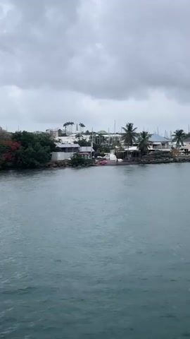 The video shows a view of a coastline from a body of water. The sky is overcast and appears to be cloudy. The coastline features several buildings, some of which appear to be houses or apartments, and others that might be part of a resort or complex. There are palm trees and other lush green vegetation along the shore. In the water, there is a small pink boat visible near the shore. Further out in the water, a sailboat with a white hull and sail is present. The water itself is a shade of blue-green and appears somewhat choppy. The camera is moving, panning from left to right, offering a continuous view of the scene.