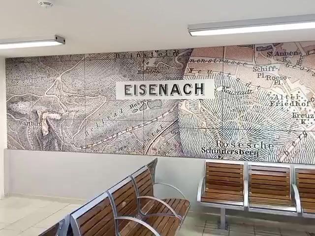 The video shows a wide shot of a waiting area. The wall behind the seating is decorated with a large, vintage-style map that says "EISENACH" in large white letters. Several rows of wooden bench seating with metal frames are visible in the foreground. The area appears to be indoors, with fluorescent lighting fixtures on the ceiling. The camera pans slightly across the scene.