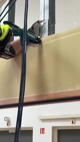 A person wearing a yellow and black glove on their left hand is using a teal and black screw gun with a strip of screws to attach a beige panel to a metal frame. The screw gun is connected to a battery pack. The person's right hand is wearing a gray and black glove and is holding the panel in place. The panel is positioned horizontally, and the metal frame is vertical behind it. There are black cables running vertically in the foreground. The background appears to be an interior space with white walls and some other fixtures visible at the bottom.