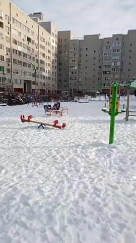 The video shows an empty, snow-covered playground located in an apartment complex. The playground is surrounded by tall, multi-story buildings.  Various playground equipment is visible, including a slide, a seesaw, and a jungle gym. A car buried in snow is visible in the background. There is no visible human activity in the playground. The sky is partially cloudy. The overall atmosphere is quiet and still.