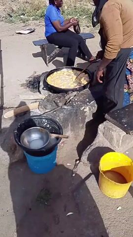 Two women are outdoors preparing food. One woman, dressed in a blue shirt and black pants, sits on a wooden bench holding a small dark object. The other woman, wearing a brown shirt and a colorful patterned apron, stands in front of a cooking setup, using a slotted spoon to move food frying in a large pan over a stone hearth. A black bucket, topped with a smaller metal bowl and a wooden stick, is next to the hearth, along with a yellow bucket. The ground is concrete, and in the background is an area of dry grass and trash.