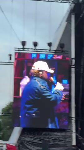 A medium shot displays a large LED screen showing a person in a denim jacket and a white baseball cap speaking into a microphone on a stage. The person is facing away from the camera, and the screen displays them from the back, slightly to the side. The person's right hand is holding the microphone to their mouth, and their left hand is gesturing. The background on the screen shows what appears to be a band with musical instruments like drums. Above the LED screen, there are several spotlights. The overall environment suggests an outdoor concert or event, with a cloudy sky visible above the stage structure.