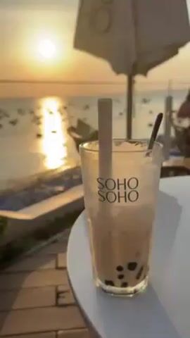 The video shows a view from a patio overlooking a beach and ocean at sunset. A clear glass with the words "SOHO SOHO" printed on it sits on a white table in the foreground. The glass contains a light brown liquid, ice, and black boba pearls. A light brown straw and a black spoon are in the glass.  A white umbrella is partially visible in the upper right corner of the frame. Beyond the table is a rattan chair and a low white structure along the patio's edge. The beach is visible with numerous small boats floating on the water, reflecting the sunlight. The sun is setting, casting a bright golden light across the water.  Two indistinct people are faintly visible in the background. A voice speaks in the background.  The focus shifts from the glass of boba to the ocean view.