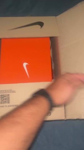 A hand reaches into an open cardboard shipping box marked with a Nike swoosh and pulls out an orange Nike shoebox. The shoebox is placed back into the larger shipping box. Then, a light-blue Nike soccer cleat with white laces and a red swoosh is lifted out of the shipping box and held for viewing. The bed has dark blue sheets, and the shipping box is held over it.