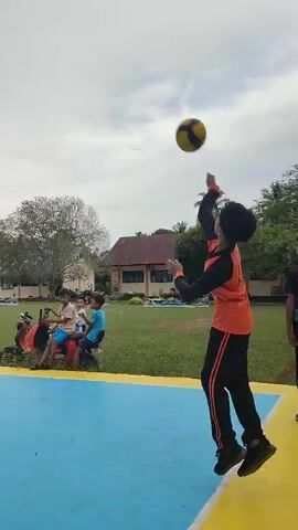 A group of young people are playing volleyball on an outdoor court. The court is marked with blue and yellow lines. There are several buildings and trees in the background. The sky is overcast. One person in an orange shirt is in the foreground, facing away from the camera, appearing to be in the middle of a serve or hit. Another person in an orange shirt is on the other side of the net. Several other people are visible on the court and on the sidelines. The ball is in the air, about to be hit.