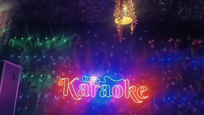 The video starts with shots of a karaoke bar, with a neon sign that says "Karaoke" and a mural that says "Music" surrounded by musical instruments and other imagery. The final shot is of a woman lying down, looking at the camera. She is wearing a white shirt and a patterned headband. The lighting is dim and colorful.