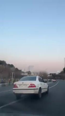 A car drives on a highway at dusk. On the driver's side of the highway there are trees and bushes, then a short barrier, then the highway.  On the other side of the barrier is another set of lanes, with cars driving in the opposite direction.  On the right is a barrier, and then a wide lane of the highway.  The sky is pale blue with hints of pink from the setting sun. There are lampposts lining the side of the highway.
