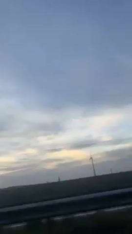 The video appears to have been taken from a moving vehicle, possibly a car or bus, looking out the passenger side window. The sky is visible, showing a mix of blue and white clouds, with some yellow hints suggesting either dawn or dusk. The landscape is mainly flat with fields and some bare trees in the background. There are also windmills visible in the distance.  A metal guardrail runs along the edge of the road. The video passes by a large grey wall or structure, briefly obscuring the view.