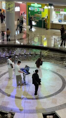 The video shows a four-person musical group performing in a mall. The group consists of a guitarist, a keyboardist, a flute player, and a person standing with a camera or other equipment. They are positioned on a circular platform surrounding a fountain in the center of the mall's open area. People are walking around the mall on different levels, and food vendors are visible in the background. The platform has lights projecting colorful patterns onto the surface. The musicians are facing away from the camera.
