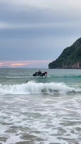 A group of people are in the ocean, some swimming and others riding a jet ski. The waves are small and choppy, and the sky is overcast with some hints of color on the horizon, suggesting either sunrise or sunset. A green, tree-covered cliffside rises from the water in the background. The water is a clear turquoise color.