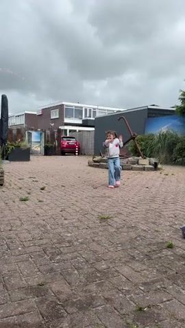 A person is running in a courtyard with a brick floor. The person is wearing blue jeans, a white sweater, and white shoes. In the background, there is a red car, a house, and a large anchor. The sky is cloudy.