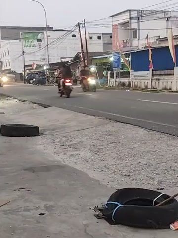 This video shows a street scene with several motorcycles visible. In the foreground, there are two discarded tires on the sidewalk. The street is paved and appears to be a commercial or urban area with buildings lining one side. The sky is overcast or it is dusk. Two motorcycles are driving on the road. One motorcycle is moving away from the camera, and the other is moving towards the camera, its headlight illuminating the road. A third motorcycle is partially visible further down the road. The overall impression is of a routine street scene with traffic and urban infrastructure.