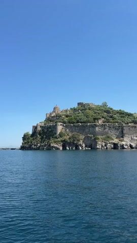 The video shows a panoramic view from a boat on the ocean. On the right side of the frame, an island with a large stone castle and green foliage sits under a clear blue sky. The castle seems ancient, constructed from stone and featuring multiple levels and a domed structure at its peak. The island's base consists of rocky cliffs that drop into the dark blue ocean water. As the camera pans slightly, a row of buoys becomes visible in the water, leading towards a town or village in the distance on the left side of the frame. The water surface has gentle ripples.
