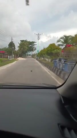 The video shows a first-person view from inside a car, looking out through the windshield. The car is driving on a paved road with two lanes. On the left side of the road, there are houses and trees, with a pile of sand. On the right side of the road, there is a sidewalk with a concrete barrier and more trees. There are utility poles and wires visible. In the distance, there are streetlights, some of which are decorated with green lights. The sky is cloudy. The road appears to be in a rural or suburban area. The video is short, with the timestamps indicating it is only a few seconds long. There are no people visible in the video, and no specific actions are taking place other than the car driving.