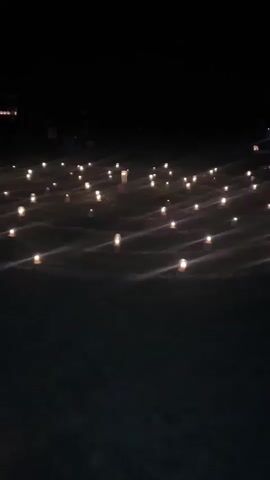 A wide shot shows a field at night, illuminated by numerous luminaries arranged in rows. The luminaries are small, cylindrical bags with a light source inside, casting a warm glow. Some luminaries appear to be taller and possibly stand on pedestals. In the far background, a few figures are visible, but they are indistinct due to the low light conditions and distance. The ground is covered in dark grass. The overall scene suggests an outdoor event or memorial.