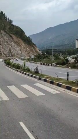 A cloudy, overcast day. A road with a pedestrian crossing in the foreground. A few cars and a truck are visible on the road. The road curves to the left, with a steep, rocky hillside on the left side. Trees and vegetation grow along the side of the road. In the distance, mountains rise up, covered in trees. The overall impression is a scenic mountain road.