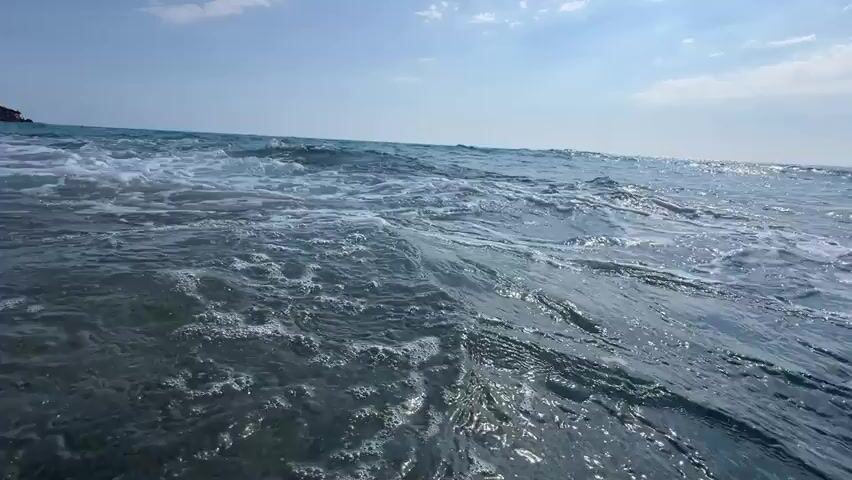 The video shows a low-angle, first-person perspective of waves breaking on a rocky shore. The water is blue and appears to be clear. The sky is blue with some white clouds. In the distance, there is a red structure, possibly a building or a pier, and a small boat or person on a jet ski can be seen in the water. The waves are small to moderate in size and are creating white foam as they crash against the rocks. The camera is submerged in the water at times, giving a unique perspective of the underwater environment.