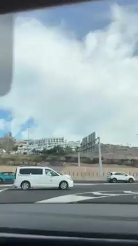 The video shows a view from a moving vehicle driving on a multi-lane road with traffic. A white van is traveling in the adjacent lane. Buildings are visible on a hill in the background. The sky is partly cloudy. The driver is being given navigation instructions.