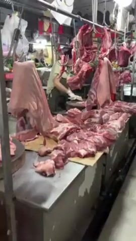 The video shows a butcher shop or market stall with a large quantity of raw pork cuts displayed on a table. A person wearing a light-colored shirt, a dark apron, and white gloves is visible in the background, cutting more meat on a wooden chopping block. Several large cuts of pork hang from hooks above the table. The table is covered with a sheet of cardboard. In the background, there is a weighing scale. The overall scene appears to be a busy marketplace. Someone asks "How much one kilo?"