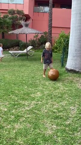 The video shows two young boys playing on a grassy lawn in front of a resort building. One boy, with blonde hair, is wearing a dark t-shirt with a graphic design and light-colored shorts. He is barefoot and positioned in the foreground, looking towards a large, brown, inflatable ball. The other boy, wearing a white t-shirt and shorts, is in the background, also barefoot, standing near a lounge chair and a straw umbrella.

The setting appears to be a resort or hotel garden, with palm trees, lush greenery, and a pink building in the background. A large tree trunk, possibly a palm tree, is visible on the right side of the frame. The ground is covered in neatly mown green grass. The lighting suggests it is daytime, likely sunny, as indicated by the bright grass and the shadows. The overall mood seems relaxed and playful, with the boys engaged in an outdoor activity. There is some text visible on the blonde boy's t-shirt, though it is partially obscured and difficult to read clearly. The location context provided is Resinera-voladilla, Spain, which aligns with the visual cues of a warm, sunny climate and resort-style environment. The activity is the boys playing with a ball.