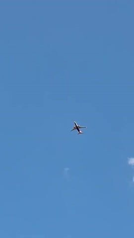 An airplane flies across a clear blue sky. A few small white clouds are visible. The plane is white with red and black markings and appears to be leaving a faint contrail.