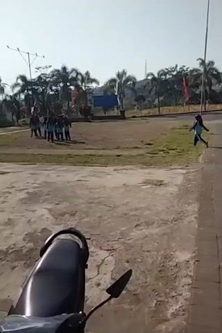 The video shows an outdoor scene with several people. In the foreground, the back of a motorcycle is visible. In the middle ground, a dirt or gravel area is present, and beyond that, a grassy field with several palm trees. There are a few people, possibly students, gathered in the left side of the grassy field. One person in a hijab and blue shirt is walking or running on the right side of the grassy field, away from the group. There are flags and a blue tarp visible in the background. The overall environment appears to be a park or an open area with some structures. The time stamp indicates the video is short and likely a short clip.