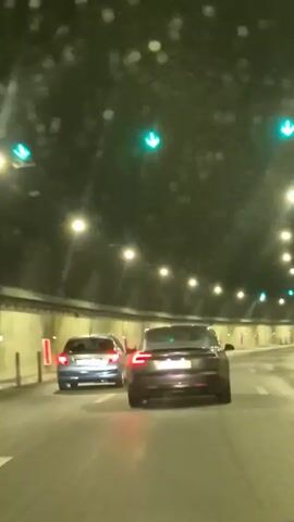 This video captures a view from inside a tunnel, likely at night or with minimal natural light. The primary subjects are two cars driving through the tunnel. The car in the foreground is a dark-colored sedan, while the car in the middle ground is a lighter-colored sedan. The tunnel is illuminated by a series of bright overhead lights, spaced regularly along its length. The tunnel walls appear to be made of concrete and are lined with vertical red and white markers. There are also several green arrow traffic signals visible on the ceiling of the tunnel, indicating that traffic flow is permitted in the direction of travel. The road surface appears to be wet, suggesting recent rain or damp conditions. The overall atmosphere is one of transit and movement through an enclosed, artificial environment. No people are clearly visible, and the focus is on the vehicles and the tunnel infrastructure. There is no visible text in the frame. The timestamp "00:00" appears in the bottom left corner of the video.