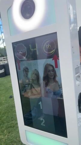 The video shows a photo booth taking pictures of three young women. The machine is white with a camera and a ring light at the top. A screen displays a countdown and instructions to "Look Up!" as it prepares to take a series of photos. The booth appears to be outdoors on a grassy area, possibly at an event or gathering, with stage structures and barriers visible in the background. Loud music plays in the background.