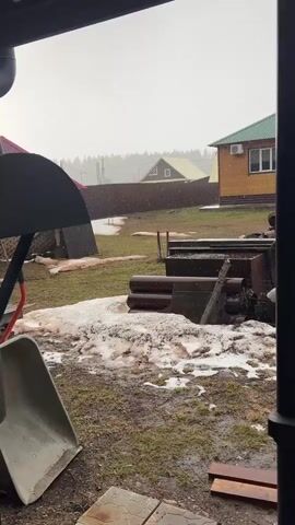 The video shows a residential backyard during a hailstorm. The ground is partially covered with melting snow and patches of grass and mud. The video is taken from under a porch or covered area, with a wheelbarrow and some metal structures visible in the foreground. Beyond the open yard, there's a wooden fence. Behind the fence are two houses, one with a cream-colored roof and another with a green roof, and a background of trees on a foggy horizon. Heavy hail is falling, visible as white specks against the gray sky. The light is dim and overcast.