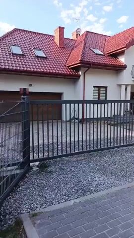 The video shows a front view of a house with a closed gate. The house is a two-story structure with a red tiled roof and white walls. It has a balcony on the upper floor with black wrought-iron railings. There are windows with blinds on the ground floor and upper floor. The entrance to the house has a covered porch with columns and a decorated door.

In front of the house, there is a dark gray metal gate, which appears to be a double gate. The gate is closed, and the two sections are latched. The ground in front of the gate is covered with small gray gravel, and a paved walkway is visible in the foreground. Beyond the gate, there is a driveway area with more gravel. To the right of the house, a mailbox is visible.

The scene appears to be set in a suburban or residential area. The sky is visible at the top of the frame and is partly cloudy. The lighting suggests it is daytime, likely late afternoon due to the shadows. The overall impression is one of a well-maintained property. No people are visible in the video. The video is a static shot, with no camera movement. The house number "8a" is visible on the wall to the right of the front door.