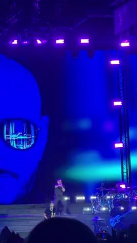 The video shows a live concert performance. A large projection of a man's face wearing sunglasses fills the background. On stage, two male performers are visible. One is singing into a microphone, standing center-stage on a raised platform. Another man is standing to the right, also on a raised platform, possibly playing a saxophone. The stage is lit with bright purple lights. In the foreground, the silhouettes of a crowd with their hands raised are visible, suggesting an audience enjoying the performance. The overall atmosphere appears energetic and dynamic, typical of a live music event.