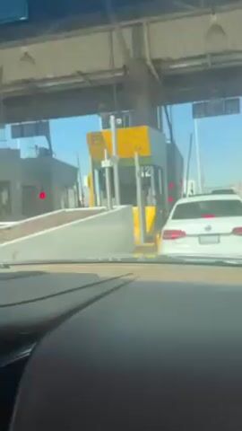 The camera is inside a car, viewing a toll booth. The toll booth has a yellow and gray exterior with the number 12 prominently displayed. A person inside the booth is taking payment from the driver of a white car. The driver extends their arm towards the booth to give the payment. The traffic light at the toll booth turns green.