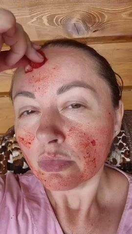 The video shows a close-up of a woman's face. She has a red substance, possibly a face mask or peel, applied to her skin, giving it a blotchy, red appearance. She is using a piece of fruit, likely a strawberry, to apply more of the red substance to her forehead and cheeks. The woman's eyes are visible, and she appears to be looking at the camera. The background shows a wooden wall and some texture that may be part of a chair or clothing.
