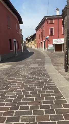 The video shows a narrow, cobblestone street in a European town. Buildings line both sides of the street, painted in various colors, including red, pink, and yellow. A street sign that reads "Mondini" with a blue arrow pointing left is visible on one of the buildings. The street curves as it leads away from the camera, with an archway visible in the distance. The sky is blue with some clouds. The sun is casting shadows on the street and buildings. There is a small white vehicle parked further up the street. The overall scene is quiet and appears to be during daytime.