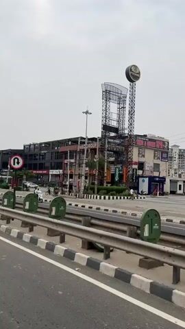A slightly overcast day in an urban environment. The video pans across a multi-story building complex with a dark facade and numerous windows. Below the complex is a street with several vehicles, including cars, a scooter, and bicycles. Green planters and a metal barrier line the foreground, separating the street from a road where traffic is flowing. The building has signage, including "SBC GROUP" and "Clarkson". Two tall, slender towers, one with a circular illuminated sign at the top, are visible to the right of the building. The overall impression is a bustling commercial area.