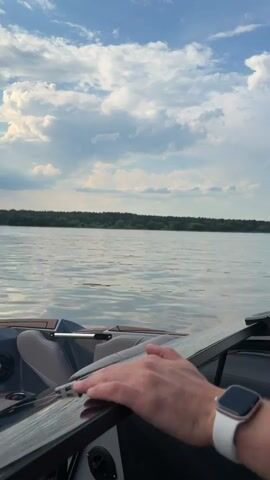 The video begins with a close-up of a person's hand on a boat's steering wheel, with a body of water and a cloudy sky visible in the background. The camera then shifts to focus on a child wearing a life jacket and a baseball cap, seated in the driver's seat of the boat and holding the steering wheel. An adult male, wearing sunglasses and a baseball cap, is seated behind the child, with his arm around the child. The child appears to be mimicking driving the boat. The video then pans away from the child and adult, showing the interior of the boat and the water outside, with a sunset visible on the horizon. The overall scene depicts a family enjoying a boat ride on a lake.