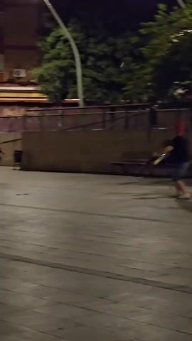 The video shows two individuals in an outdoor area at night. The ground is paved with wood. In the background, there is a raised concrete structure with a wooden bench. Trees are visible behind the structure. A street lamp illuminates the scene. One person, wearing a red cap, dark t-shirt, and shorts, is standing and appears to be interacting with a second person who is seated on the bench. The second person is mostly obscured. The person in the red cap is in motion, facing the bench.