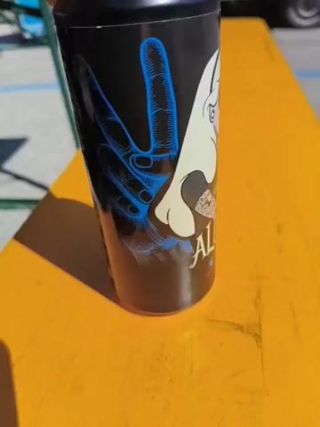 The video shows a black insulated mug with an image of a tattooed, sunglasses-wearing Alice from Alice in Wonderland design on a bright yellow table. The image is centered on the side of the mug, with a large blue hand forming a peace sign partially visible behind her. The text "Alice 5.0" is displayed beneath the illustration. The table appears to be outdoors and the camera slowly rotates the mug to show the design more completely. There are a few other items visible in the background, including the legs of a green chair and a parked car in the distance.