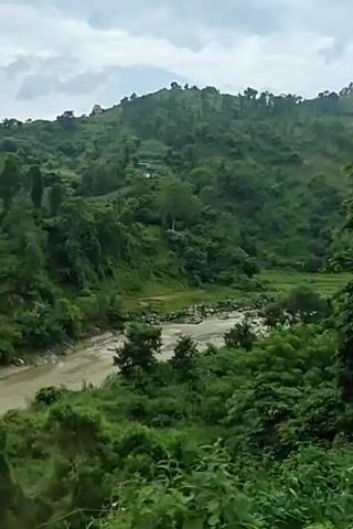 A drone shot moves forward over a lush green landscape. The scene is dominated by rolling hills covered in dense vegetation. A muddy river flows through a valley in the center of the frame, with large rocks visible in its bed. Trees and foliage line the riverbanks and cover the slopes of the hills. In the distance, a few buildings are visible nestled on the hillsides. The sky is overcast, suggesting a cloudy day. The camera movement is smooth, providing a sweeping view of the natural environment.