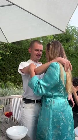 The video shows a man in a white t-shirt and light jeans hugging a woman in a teal robe with floral embroidery on a patio. A partially obscured man is sitting at the table. There are drinks and a bowl on the table. The camera pans to a backyard featuring a pool, lush landscaping, and people sitting and standing in groups. The atmosphere appears relaxed and casual.