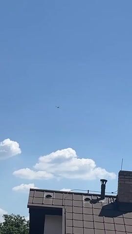 The media file captures a clear, sunny day with a bright blue sky. A small, dark silhouette of what appears to be a helicopter or small aircraft is visible in the upper center of the frame, navigating the vast expanse of the sky. Below the aircraft, fluffy white clouds drift gently, adding depth and texture to the aerial view. The foreground is dominated by the roof of a building, tiled with dark brown shingles. A brick chimney stands prominently on the right side of the roof, topped with a black ventilation cap. The sunlight casts distinct shadows, indicating it is likely midday. The overall scene suggests a peaceful, everyday moment, possibly observed from a window or balcony in a residential area. There is no visible text within the frame.
