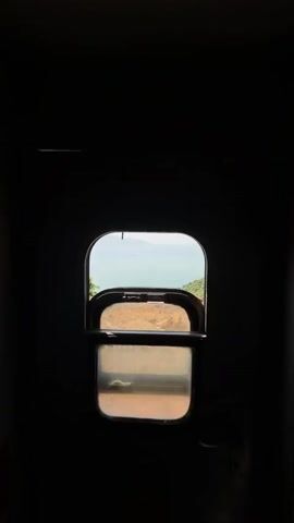 The camera looks out of a window of a moving train. The initial view is a close up of the window, which opens up to reveal a lush green hillside that slopes down to the ocean. The ocean is calm and blue. The sky is clear, with a few wispy clouds. As the train moves, the view from the window shows the hillside and ocean at eye level. There are utility poles with multiple lines of wire in the sky above. The hillside is a mix of dirt, rocks, and greenery.