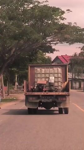 A daytime video clip from Matangpineung, Indonesia, shows the rear view of a rusty, light-brown dump truck or flatbed truck with high sides, driving down a paved road. The truck is carrying a large, clear, rectangular intermediate bulk container (IBC) tote with a metal cage around it, positioned prominently in the center of the bed. Several other smaller, dark-colored objects, possibly machinery or engines, are also visible in the truck bed, along with what appears to be some white sacks or covers.

A man on a white scooter, wearing a plaid shirt and flip-flops, follows closely behind the truck. The road features a yellow dividing line. Lush green trees line both sides of the road, and in the background, several houses with red-tiled roofs are visible, suggesting a rural or semi-urban residential area. Towards the end of the clip, another scooter rider, wearing a helmet, passes the truck and the following scooter from the opposite direction. The sky is bright, indicating clear weather. No discernible text is clearly visible on the vehicles.