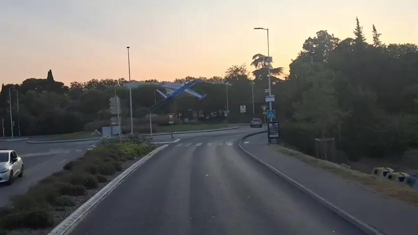 This video captures a drone shot of a roundabout in Salon-de-Provence, France, at dusk. The sky is a soft gradient of orange and purple, indicating the setting sun. A prominent sculpture of a blue and white jet airplane, tilted as if in mid-flight, is the centerpiece of the roundabout. Trees and lush greenery surround the area, creating a natural backdrop. Road signs are visible, including yield signs and directional signs, suggesting this is a junction leading into or out of the town. A single car can be seen driving on the roundabout. The overall atmosphere is serene and picturesque, highlighting the unique artistic installation within its urban environment. The time of day and clear skies suggest pleasant weather.
