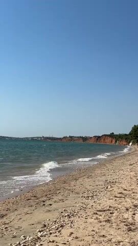 The video shows a wide shot of a beach with a shoreline and the ocean. The beach is covered in sand and pebbles. Small waves are rolling onto the shore. In the background, there is a red cliff face that curves along the coastline, and beyond that, there are some buildings and trees. The sky is clear and blue. The video is taken from a stationary perspective and there are no people or animals visible. The lighting suggests it is daytime, likely sunny.