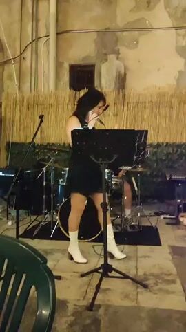 A woman with dark hair, wearing a black dress and white boots, sings into a microphone on a stage. She stands in front of a music stand, with a drum set behind her. The stage appears to be outdoors, with a textured wall and bamboo matting in the background.
